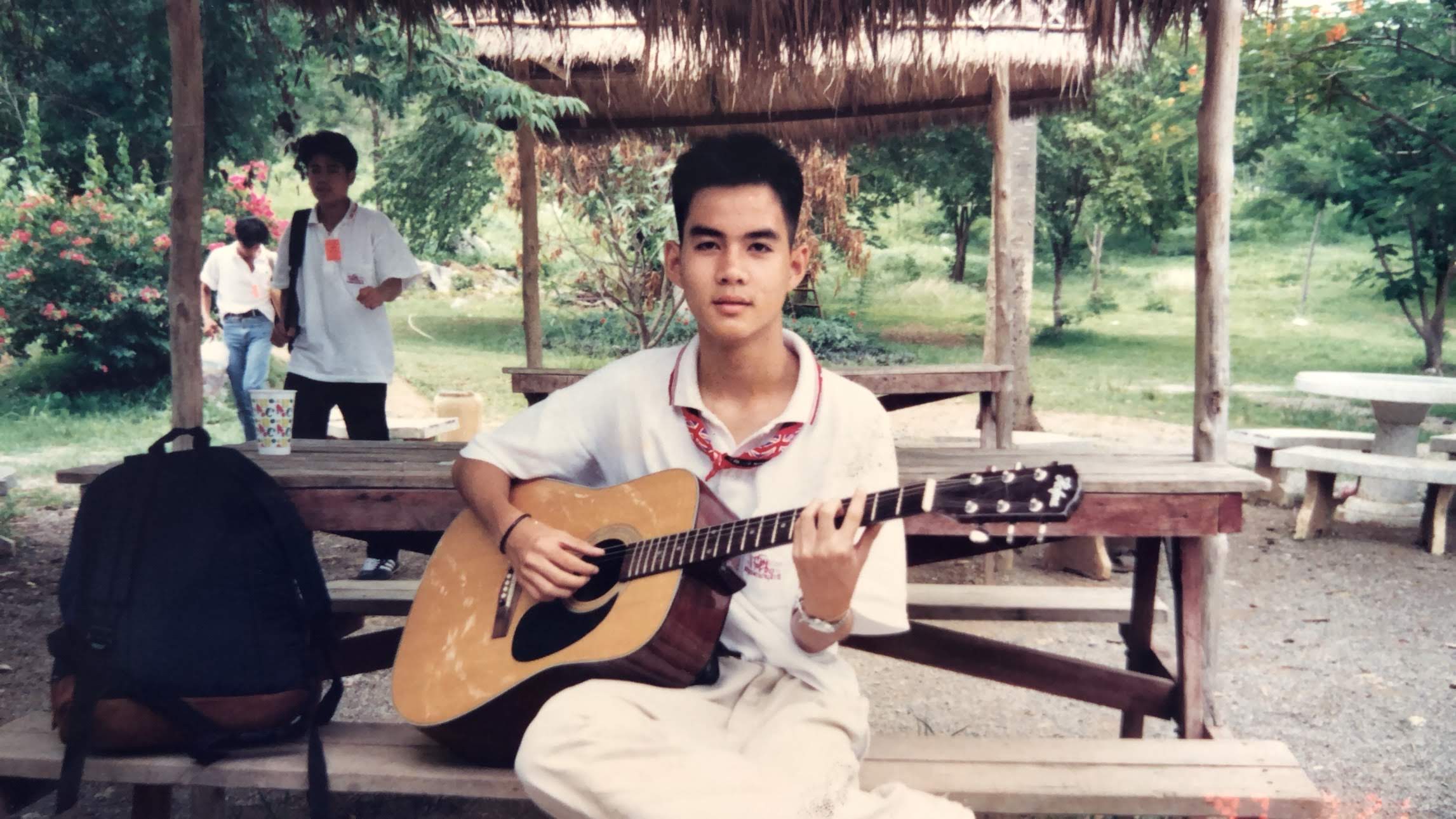 Non in school uniform playing guitar in an outdoor pavilion, Thailand — before the titles
